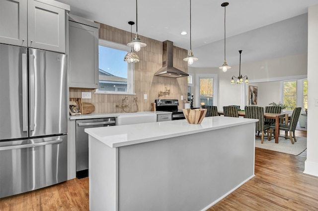 kitchen with hanging light fixtures, stainless steel appliances, wall chimney range hood, and light hardwood / wood-style floors