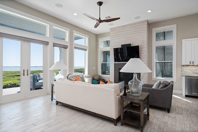 living room with a water view, ceiling fan, a large fireplace, light hardwood / wood-style flooring, and french doors