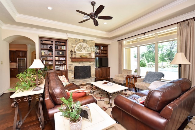 living room with ceiling fan, a stone fireplace, dark wood-type flooring, built in features, and ornamental molding