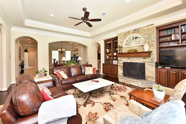 living room featuring built in features, ceiling fan with notable chandelier, crown molding, a tray ceiling, and a stone fireplace