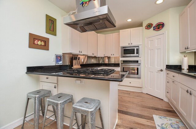 kitchen featuring appliances with stainless steel finishes, a breakfast bar, light wood-type flooring, white cabinetry, and island exhaust hood