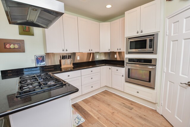 kitchen with appliances with stainless steel finishes, white cabinets, light wood-type flooring, extractor fan, and dark stone countertops