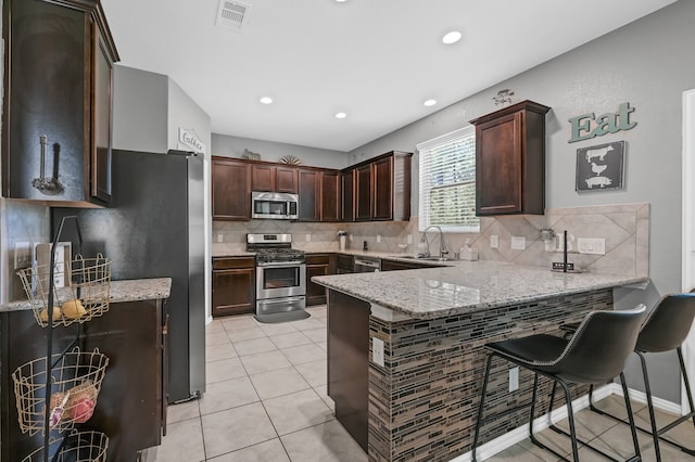 kitchen with light stone countertops, backsplash, stainless steel appliances, a kitchen bar, and light tile floors