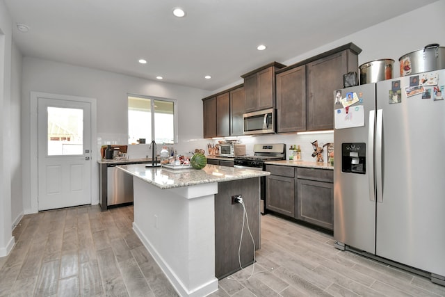 kitchen with dark brown cabinetry, a center island, stainless steel appliances, light stone counters, and light wood-type flooring