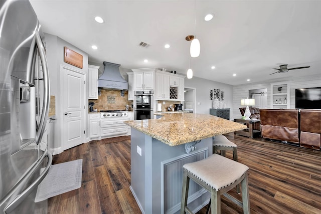 kitchen with appliances with stainless steel finishes, hanging light fixtures, custom range hood, dark hardwood / wood-style floors, and a breakfast bar