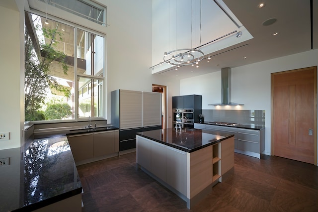 kitchen with dark tile flooring, dark stone counters, wall chimney range hood, an island with sink, and sink