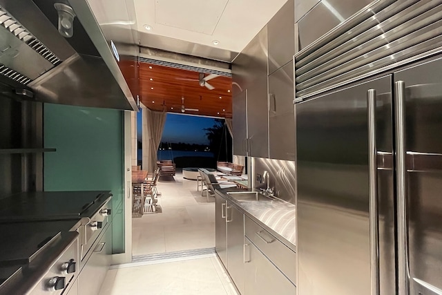 kitchen with built in fridge, stainless steel counters, wall chimney exhaust hood, sink, and light tile floors