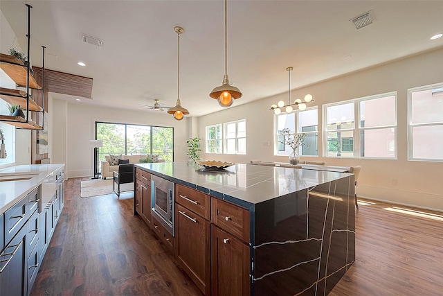 kitchen featuring ceiling fan, stainless steel microwave, dark wood-type flooring, dark brown cabinets, and a kitchen island