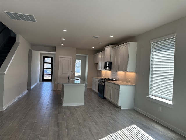 kitchen featuring sink, gas stove, a center island with sink, light stone countertops, and backsplash