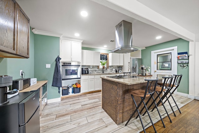 kitchen with appliances with stainless steel finishes, white cabinetry, a center island, light stone counters, and island range hood