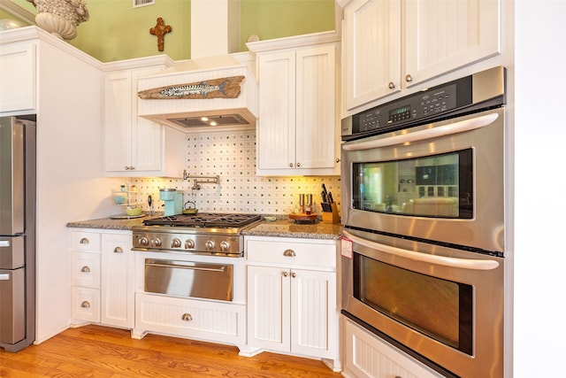 kitchen featuring custom exhaust hood, backsplash, appliances with stainless steel finishes, light wood-style floors, and light stone countertops