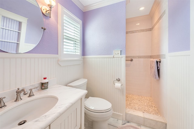 bathroom with a wainscoted wall, toilet, ornamental molding, a tile shower, and vanity