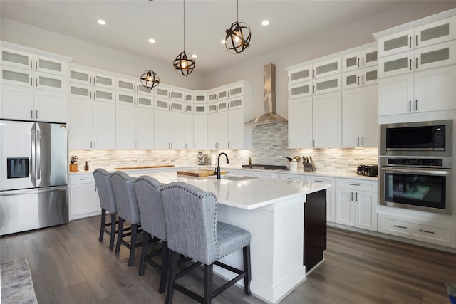 kitchen featuring a center island with sink, stainless steel appliances, dark wood-type flooring, a sink, and wall chimney range hood