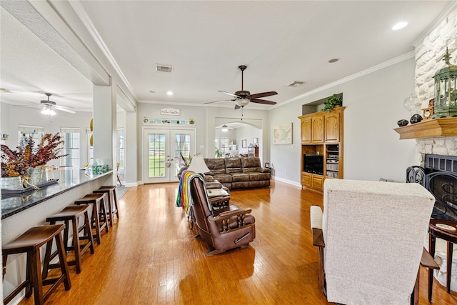 living room with ceiling fan, ornamental molding, french doors, a stone fireplace, and light hardwood / wood-style floors