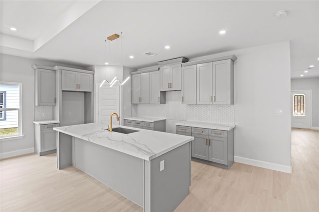 kitchen with a kitchen island with sink, gray cabinets, light hardwood / wood-style flooring, sink, and tasteful backsplash