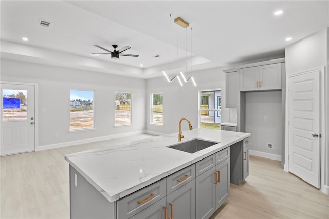 kitchen featuring ceiling fan, a tray ceiling, a kitchen island with sink, sink, and light wood-type flooring