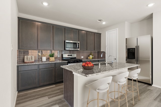 kitchen with dark brown cabinetry, a kitchen island with sink, stainless steel appliances, and backsplash