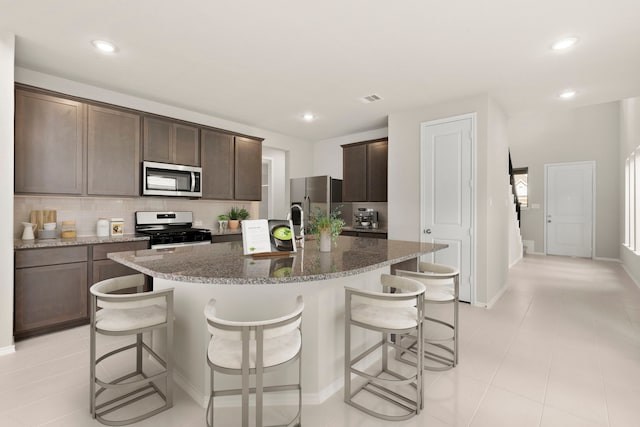 kitchen with tasteful backsplash, stainless steel appliances, dark stone counters, and an island with sink