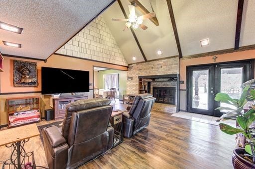 living room with a brick fireplace, ceiling fan, a textured ceiling, hardwood / wood-style flooring, and beamed ceiling