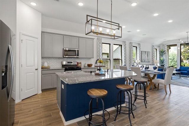 kitchen with tasteful backsplash, appliances with stainless steel finishes, light stone counters, a kitchen island with sink, and gray cabinets