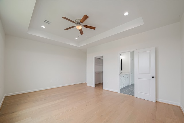 unfurnished bedroom featuring ceiling fan, ensuite bathroom, a tray ceiling, and light wood-type flooring