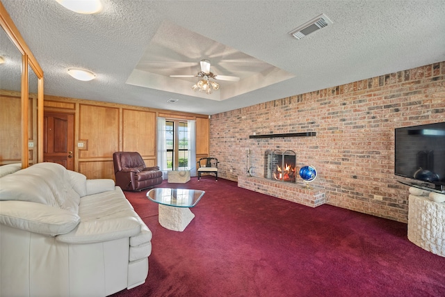 living room with ceiling fan, carpet flooring, a brick fireplace, a raised ceiling, and a textured ceiling