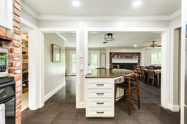 kitchen with a kitchen breakfast bar, ceiling fan, a fireplace, dark tile flooring, and white cabinets