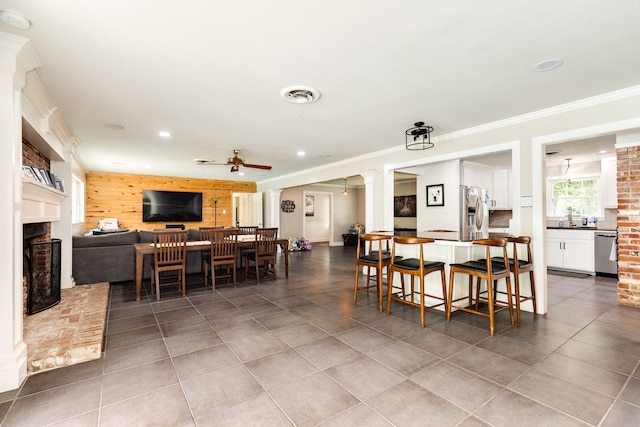 tiled dining area featuring brick wall, a fireplace, ceiling fan, ornamental molding, and ornate columns