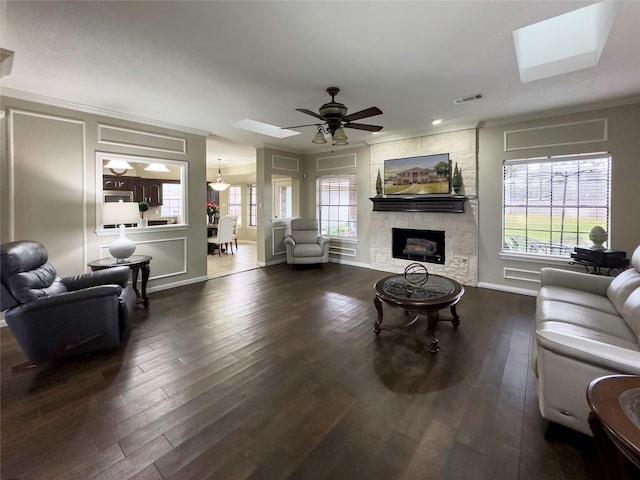 living room with crown molding, dark hardwood / wood-style floors, a stone fireplace, ceiling fan, and a skylight