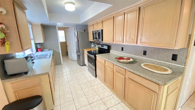 kitchen with a tray ceiling, stainless steel appliances, light tile flooring, light brown cabinetry, and backsplash