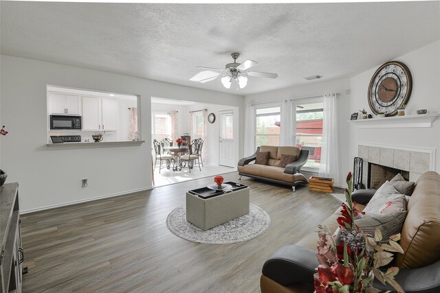 living room featuring ceiling fan, a textured ceiling, a tiled fireplace, baseboards, and wood finished floors