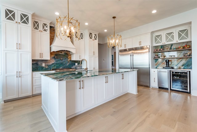 kitchen with light wood-style flooring, beverage cooler, premium range hood, stainless steel built in fridge, and an island with sink