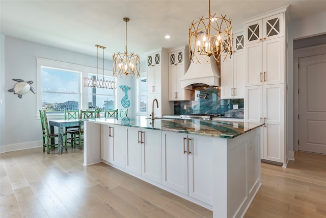 kitchen featuring a kitchen island with sink, light wood-style flooring, a sink, backsplash, and glass insert cabinets