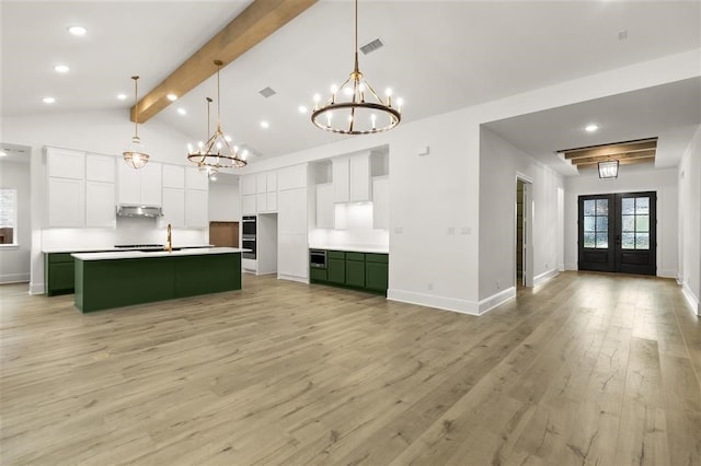 kitchen with beamed ceiling, hanging light fixtures, green cabinets, a center island with sink, and light wood-type flooring