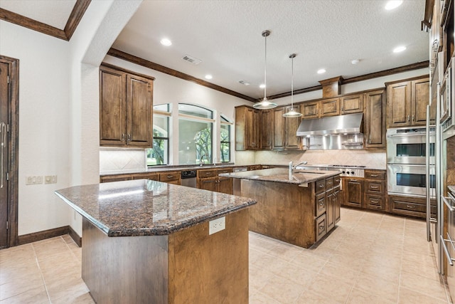 kitchen featuring a kitchen island with sink, dark stone countertops, decorative backsplash, decorative light fixtures, and stainless steel double oven