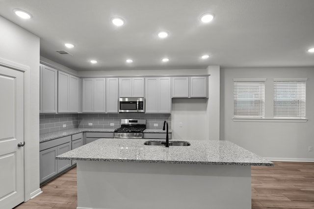 kitchen with a kitchen island with sink, light stone counters, sink, appliances with stainless steel finishes, and light hardwood / wood-style floors