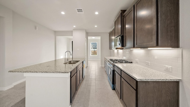 kitchen featuring appliances with stainless steel finishes, light stone counters, dark brown cabinets, sink, and a center island with sink