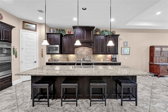 kitchen featuring dark brown cabinetry, stainless steel appliances, a large island with sink, hanging light fixtures, and a breakfast bar area