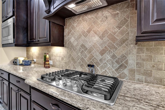 kitchen featuring light stone countertops, backsplash, premium range hood, dark brown cabinets, and appliances with stainless steel finishes