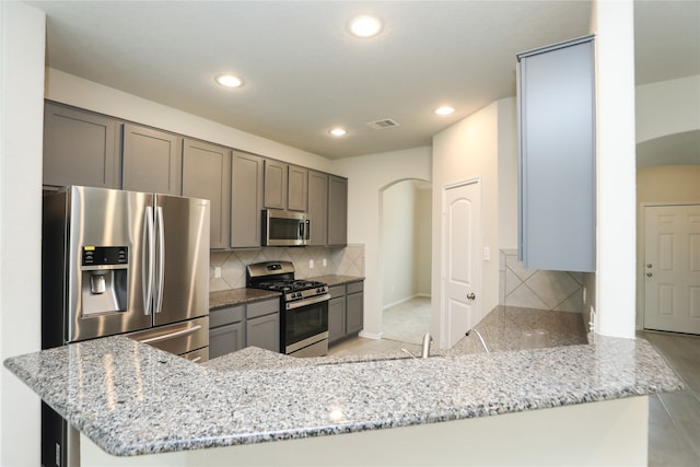 kitchen with kitchen peninsula, decorative backsplash, light stone countertops, gray cabinetry, and stainless steel appliances