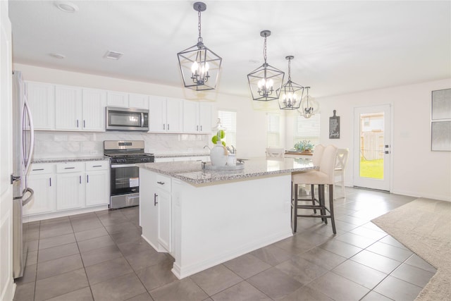 kitchen with hanging light fixtures, an island with sink, stainless steel appliances, light stone countertops, and white cabinets