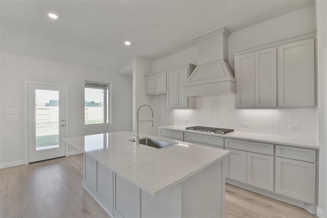 kitchen featuring stainless steel gas cooktop, custom exhaust hood, an island with sink, and light stone counters