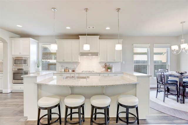 kitchen featuring backsplash, stainless steel appliances, decorative light fixtures, a center island with sink, and white cabinets