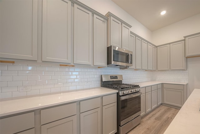 kitchen with gray cabinetry, light stone countertops, backsplash, appliances with stainless steel finishes, and light wood-type flooring