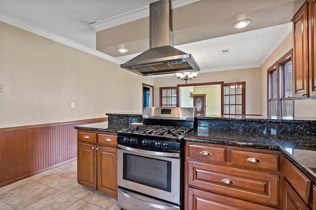 kitchen featuring island exhaust hood, dark stone counters, wood walls, ornamental molding, and stainless steel gas range oven