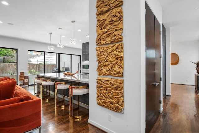 kitchen featuring dark wood-type flooring, a breakfast bar, stainless steel oven, hanging light fixtures, and light stone countertops