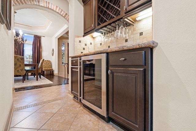 bar with light stone countertops, dark brown cabinets, beverage cooler, crown molding, and light tile patterned flooring