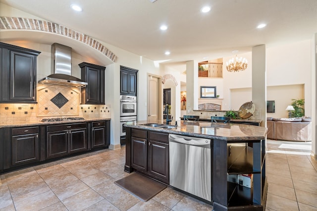 kitchen featuring wall chimney range hood, sink, an island with sink, a notable chandelier, and stainless steel appliances