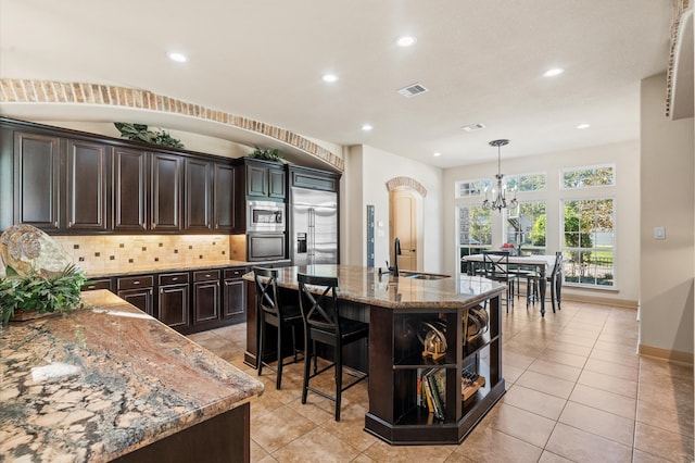 kitchen with sink, light stone counters, built in appliances, an island with sink, and a chandelier