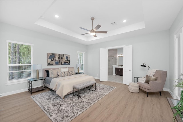 bedroom with ensuite bath, light hardwood / wood-style flooring, a raised ceiling, and ceiling fan